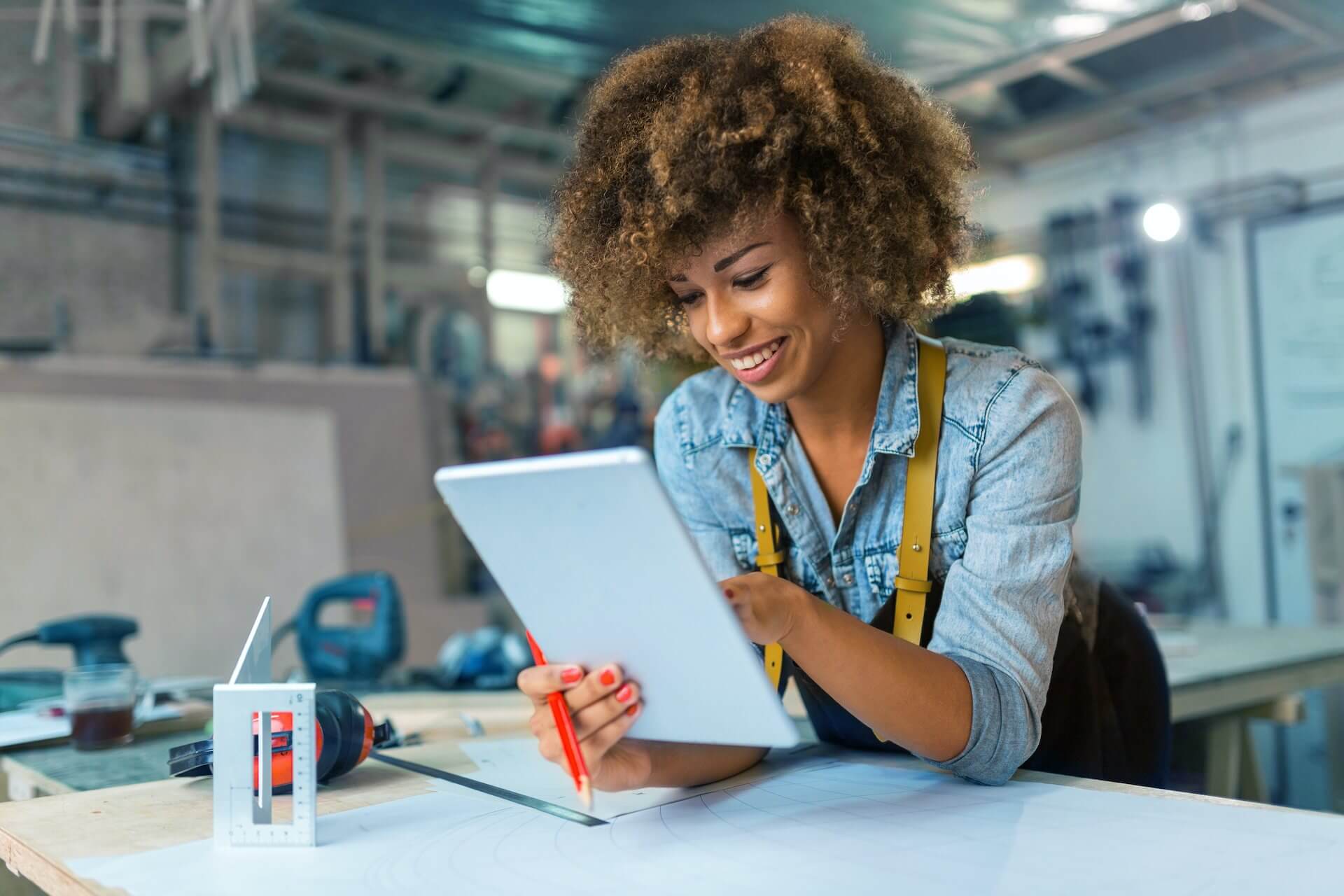 Smiling Black woman wearing apron and leaning on table to looking at iPad in studio