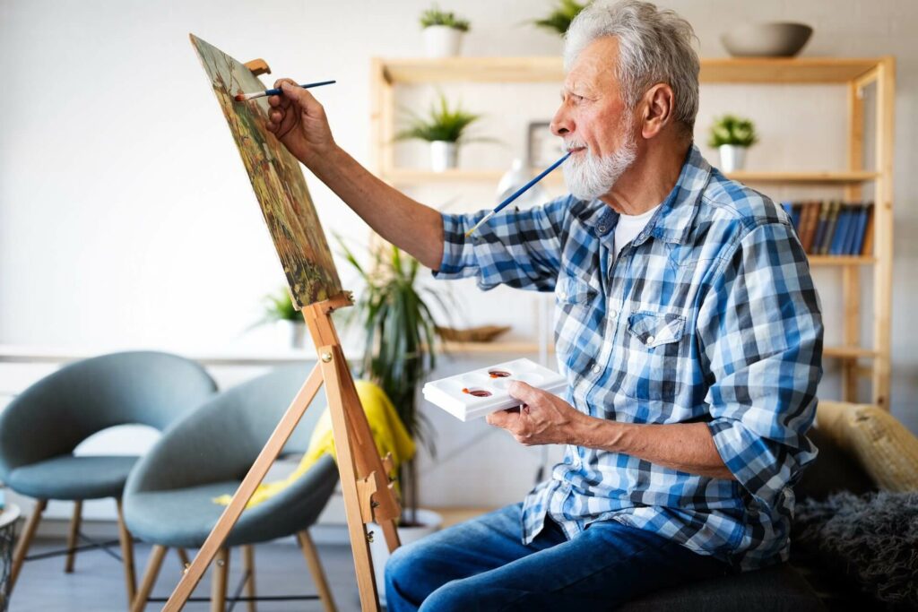 Older white man wearing blue plaid shirt painting on a wooden panel at an easel