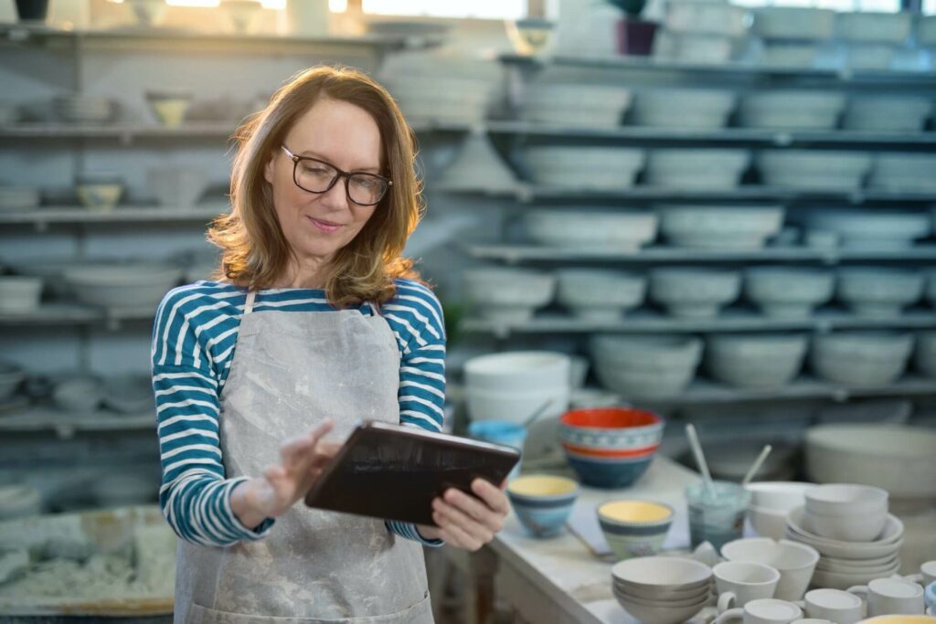 Smiling White woman with glasses working in iPad in front of dozens of clay pots