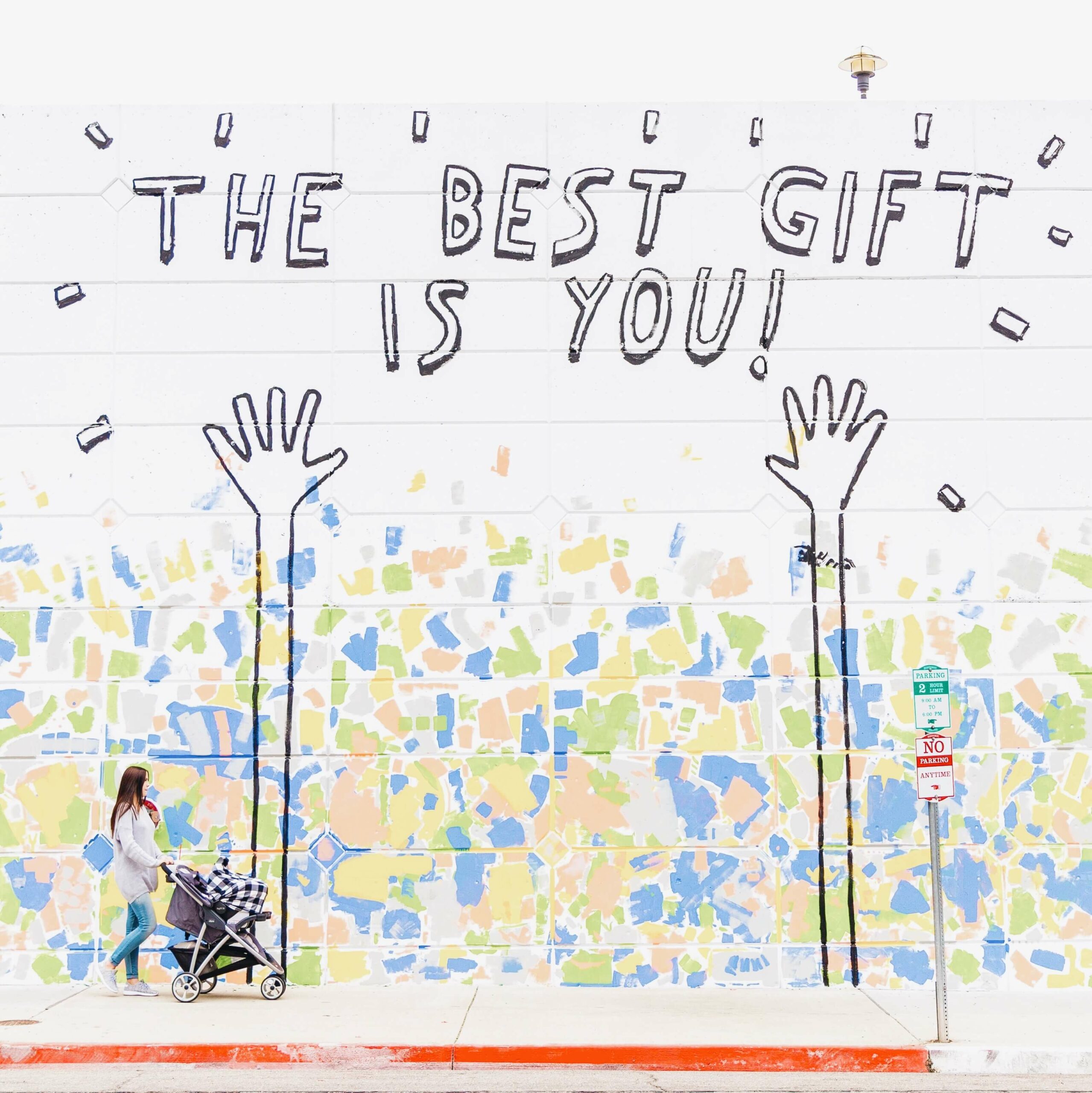 Woman with stroller walking past wall with the text "The Best Gift is You" with hands and multi-colored confetti