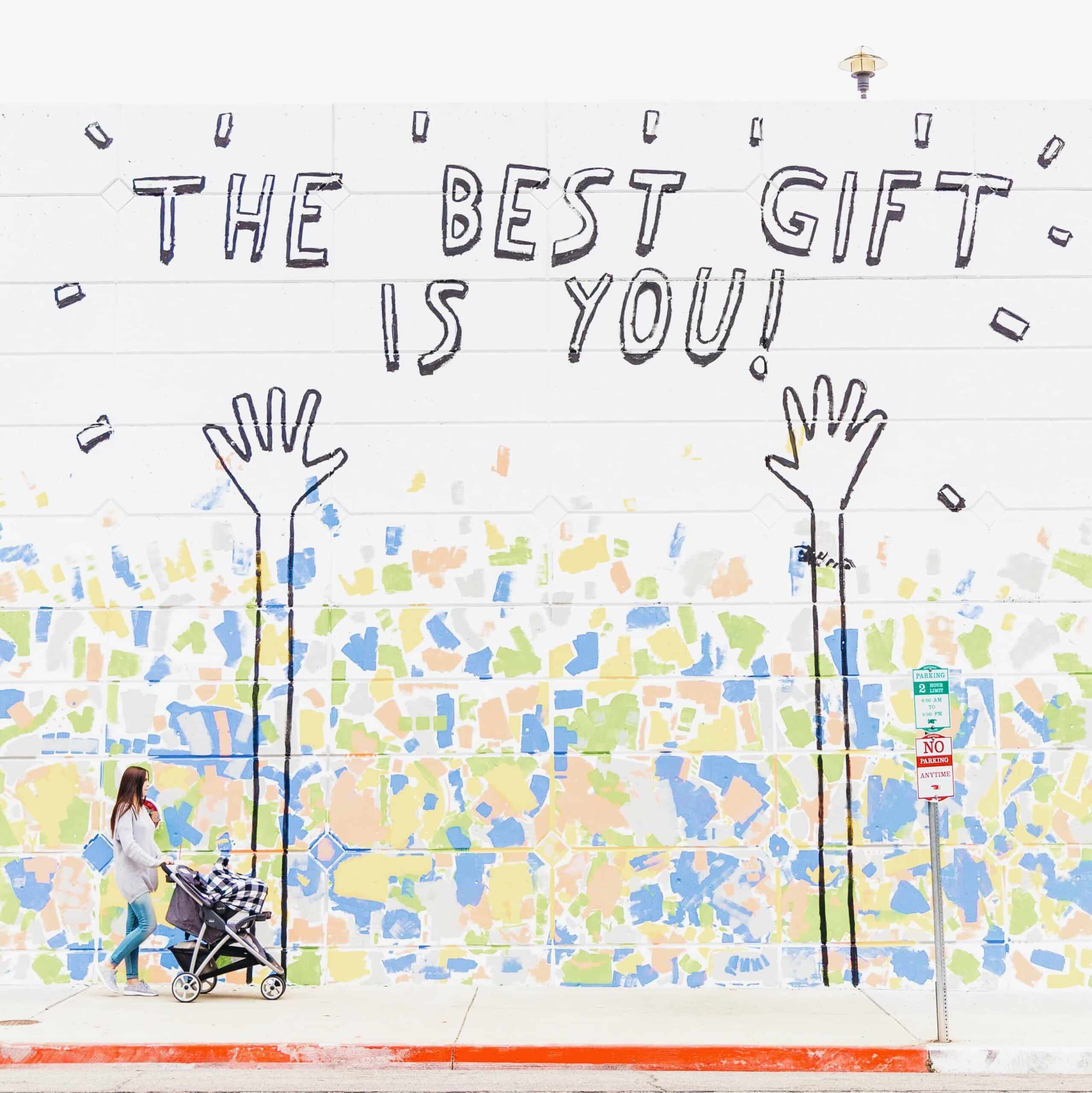 Woman with stroller walking past wall with the text "The Best Gift is You" with hands and multi-colored confetti